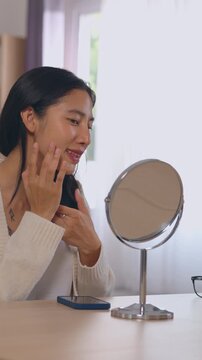 Young adult Asian woman examines her face and skin reflection in mirror, touching her cheek during skincare routine in softly lit bedroom with natural light and calm beauty focused expression