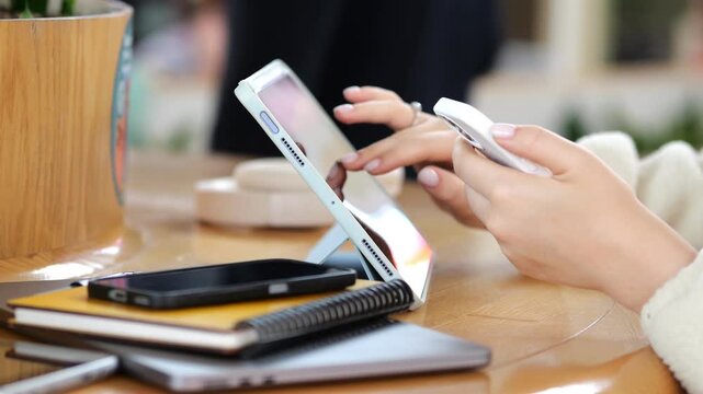 Hands using tablet and phone on wooden table. Multiple devices nearby. Blurred background with people. Coffee cup and notebook visible