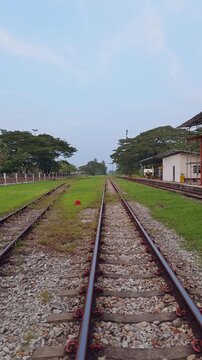 Vertical video. Parallel Railway Tracks Extend Into the Distance Beside a Station Platform and White Building, With Gravel, Green Grass and Trees Beneath a Pale Blue Sky, Showing Transport Infrastruct