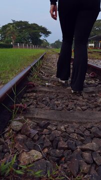 Vertical video. Woman with a Ponytail Walking Away Along Railway Tracks in Black Clothing, Low Angle Rear View Showing Gravel, Sleepers, Green Grass and Trees Near a Station