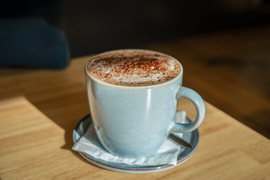 A pastel-blue ceramic cup of frothy cappuccino, delicately dusted with chocolate, basks in warm sunlight against the softly blurred shadows of a cosy caf&eacute;, Victoria, Australia
