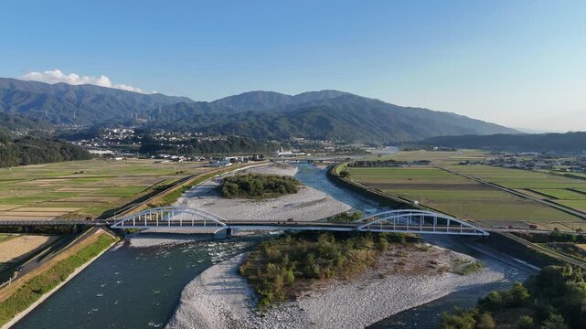 Aerial footage shows two white bridge arches over a winding river and gravel deposits amid a rural agricultural valley.