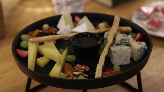 Cheese platter on dark plate with grapes, sliced apples, breadsticks, walnuts, honey dip, elegant catering appetizer for restaurant table and wine pairing.
