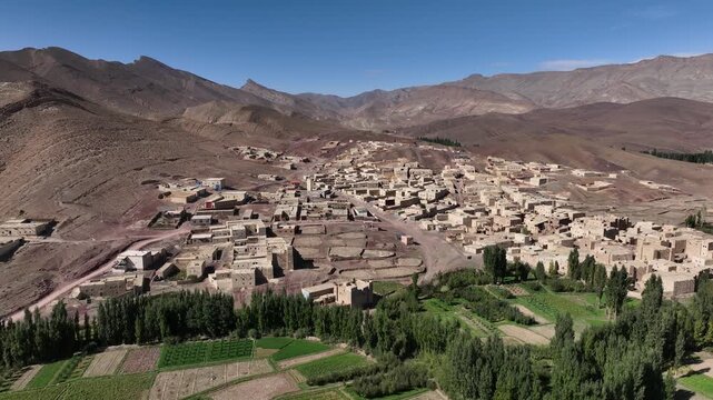Wide aerial view of a traditional mountain village surrounded by green agricultural plots, trees and rugged arid mountains in Morocco's Eastern High Atlas.