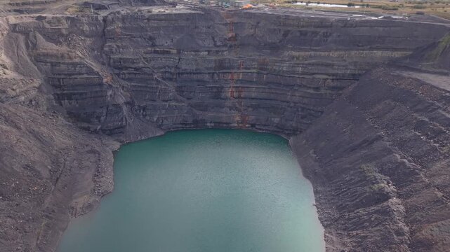 Drone pulls backward from a turquoise quarry lake, gradually revealing the massive terraced rock walls and scale of a deep open pit quarry in Wales. Dramatic industrial landscape shaped by mineral ext