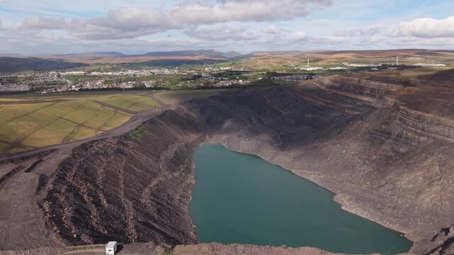 Forward aerial flight over the edge of a vast open pit quarry in Wales, revealing a deep turquoise lake below, steep excavated slopes and layered rock formations. The movement creates a dramatic revea