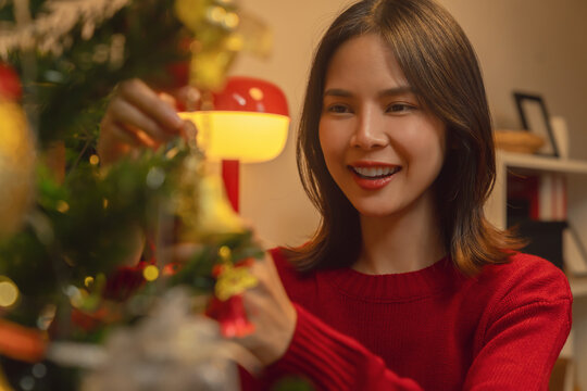 Cheerful Asian woman wearing a red sweater, decorating a Christmas tree with a festive ribbon and ornaments, holiday celebration.