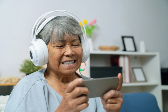 Elderly Asian woman relaxing on a sofa at home, wearing headphones while enjoying games on her smartphone, modern living among older adults.