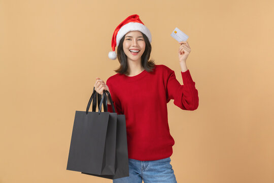 Cheerful Asian woman celebrating Christmas wearing a santa hat and holding a credit card with shopping bag while smiling, black friday online shopping and modern lifestyle concept.