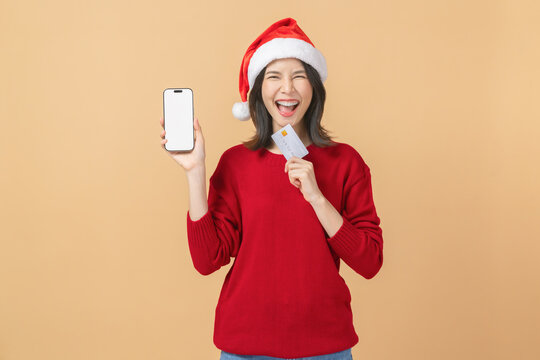 Cheerful Asian woman celebrating Christmas wearing a santa hat and holding a smartphone and credit card while smiling, online shopping and modern lifestyle concept.
