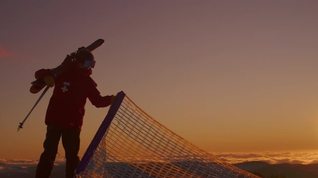 Ski patroller carries skis and holds mesh safety fencing against an orange mountain sky