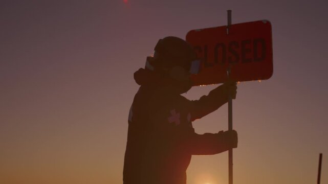 Ski patroller holds a closed sign on a pole against a glowing orange sky in low golden sunlight