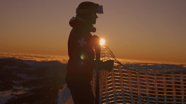 Ski patroller holds mesh safety fencing beside snowy mountain slopes beneath an orange sky