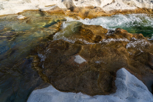 High-angle view of a mountain stream cascading around sculpted stone slabs with warm amber tones and pale chalky edges. Crystal-clear pools and frothy ripples show the power of moving freshwater