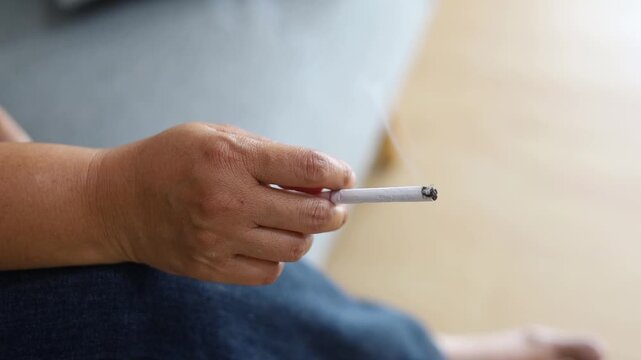 A close-up view of a person's hand holding a lighted cigarette with smoke rising, depicting smoking, tobacco use, addiction, bad habits, and health concerns.
