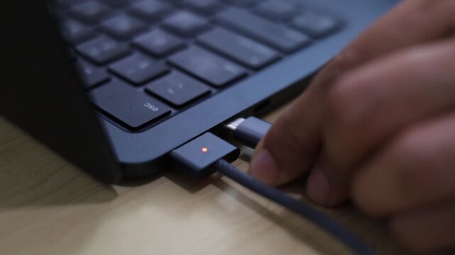 A close-up view of a person plugging a charging cable into a laptop port, depicting technology, power supply, device connectivity, and work-from-home routine.