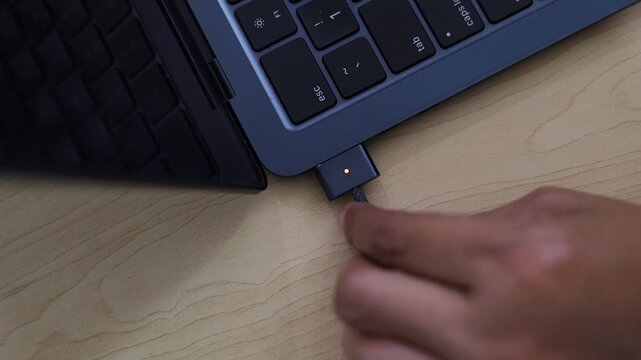 A top angle close up view of a person connecting or unplugging a magnetic charging cable to a modern laptop, depicting technology, power supply, connectivity, and workspace routine.