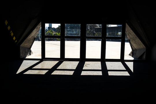 Dramatic bright sunlight streaming through grid glass doors creates strong geometric shadows and contrast on the floor of a dark concrete building interior, looking out towards