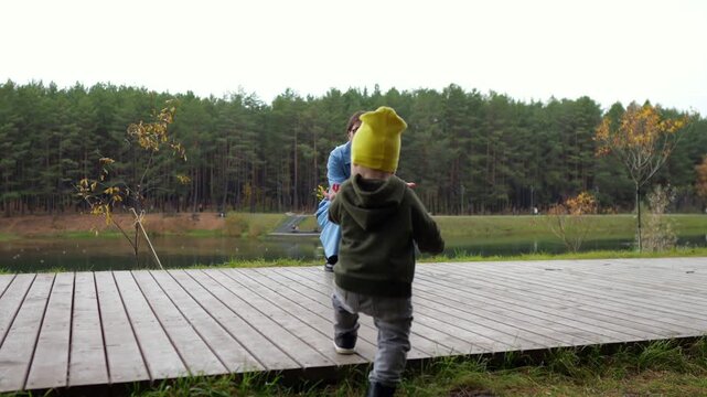 Toddler Runs Towards Mother On Wooden Pier, Stumbling Into Clumsy Fall While Approaching Open Water, Presenting Serious Child Safety Risk During Unsupervised Outdoor Family Recreation In Park.
