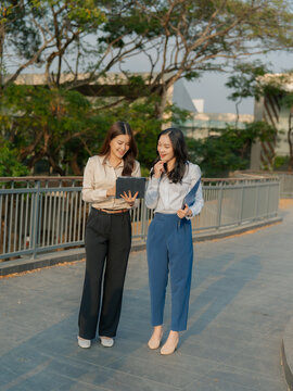 Two Asian businesswomen are discussing a work project while holding documents and a tablet outdoors, near an office building.