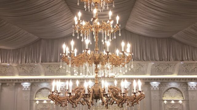 Drone orbit shot of a golden crystal chandelier with candle-style bulbs hanging beneath a draped ceiling in a decorated banquet hall with additional chandeliers and classical wall moldings.