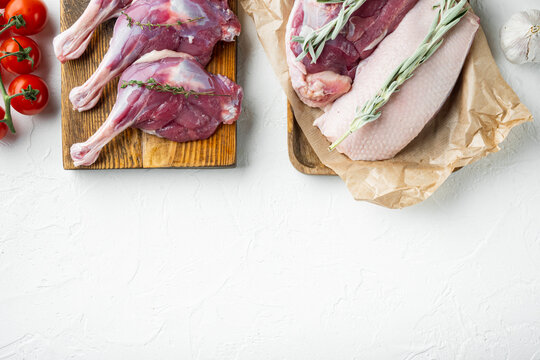 Raw duck leg thigh and breast fillet on wooden boards with fresh thyme sprigs cherry tomatoes and garlic bulb on white textured surface top down flat lay composition