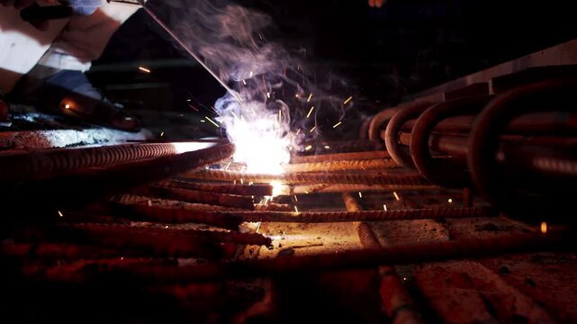 Low-light wide shot of an industrial welder performing electric arc welding on heavy steel structural rebar frameworks, creating a bright flare and showers of glowing sparks outdoors