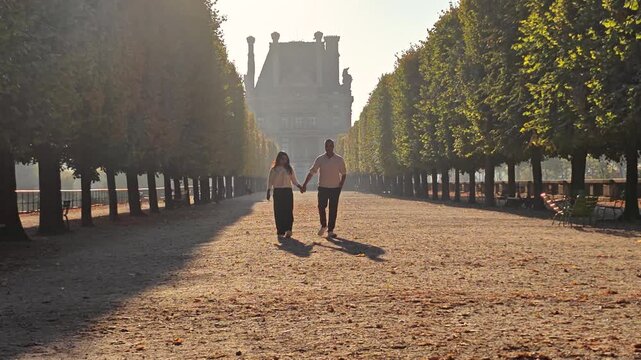 A couple walks hand in hand along the tree-lined paths of Tuileries Garden in Paris, France. The early morning sun creates a warm glow, enhancing the romantic ambiance.