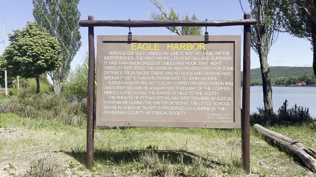 A brown historical marker beside Eagle Harbor recounts the visit of Horace Greeley copper mining settlement and Justus Rathbone with trees and Lake Superior in the background.