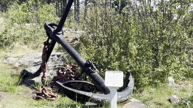 The view moves closer to a black iron stock anchor and heavy reddish chain at Eagle Harbor Lighthouse in Michigan with an interpretive placard rocks grass and green shrubs.