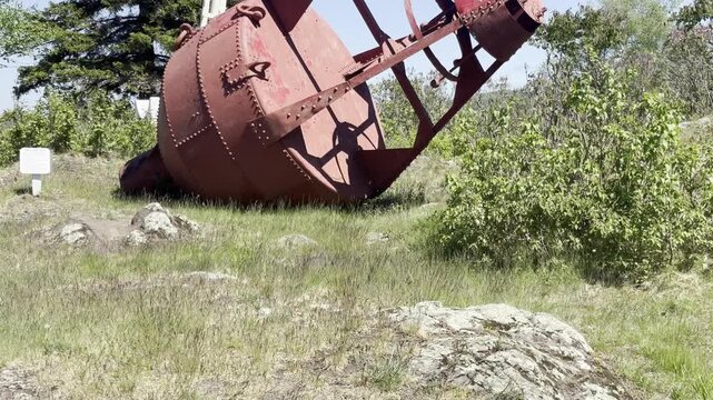 A closer view shows a red riveted bell buoy resting on rocky grassy ground at Eagle Harbor Lighthouse in Michigan with metal framework green shrubs and an interpretive sign.