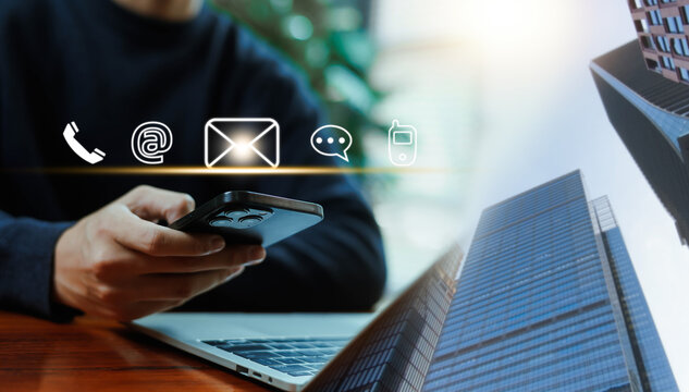 Person using a smartphone with glowing contact icons including phone, email, and chat over a laptop and city background. Concept of customer support, business communication, and connection.