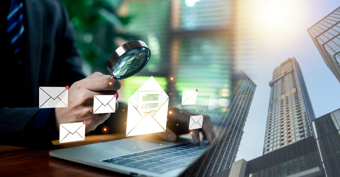 Business person holding a magnifying glass examining digital email icons and notifications over a laptop and city background. Concept of email security, message auditing, and corporate communication.