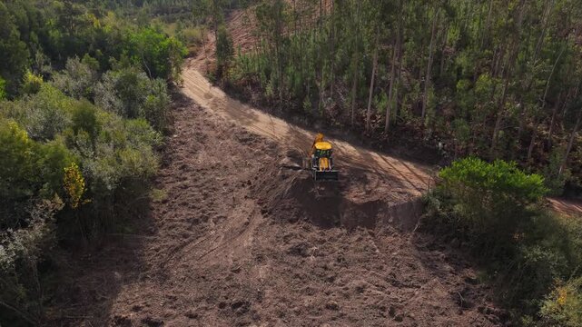 Drone shot of a yellow backhoe loader cutting a dirt track through eucalyptus woods and scrub on a hillside, part of a high speed railway project in Portugal.
