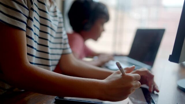 Focused creative student drawing on a digital tablet with a stylus while another young boy is doing his homework on a laptop computer in the background at home