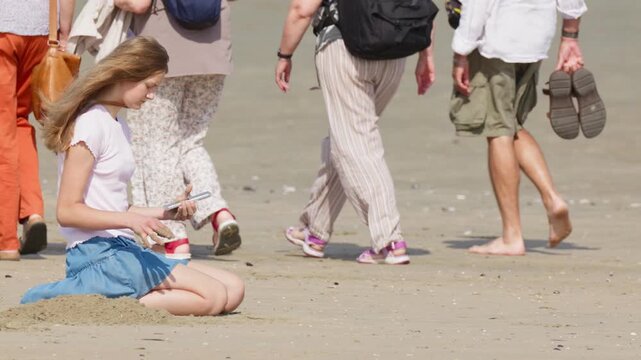 A girl buries her smartphone in the sand digs a hole with her hands a group of unrecognizable people is walking by