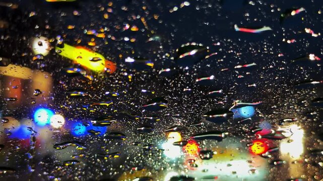 View from inside a moving car through a rain-soaked windscreen at night, with blurred streetlights and glowing light trails creating an atmospheric night time driving scene