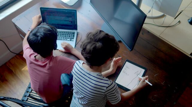 Two young brothers studying together at a desk, one boy is programming on a laptop with headphones while the other draws on a digital tablet with a stylus, representing modern e-learning