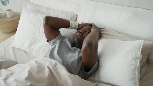 Exhausted african man lying in bed after sleepless night, portrait in morning. Insomnia and depression, disorder of mental health, intrusive thoughts, panic attack and headache, suffering male person