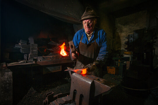 Senior blacksmith forging glowing red hot iron on anvil with hammer in traditional rustic workshop