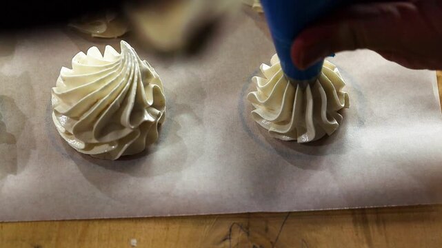 Baker's hand with blue piping bag creates decorative meringue peaks on parchment paper, showcasing the intricate design process in a well-lit kitchen environment