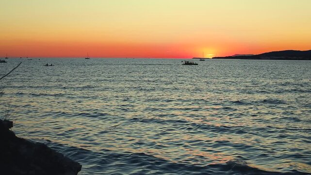 Sunset over calm water with boats sailing, silhouetted trees framing the scene, and distant hills creating a serene landscape in a coastal region