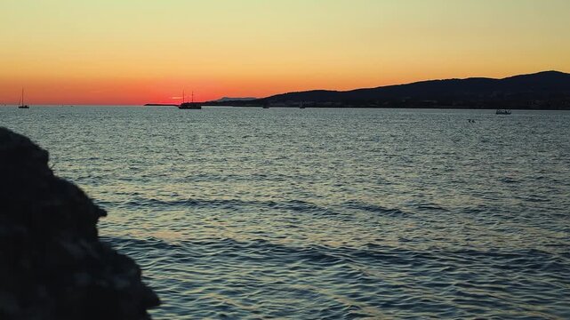 Sunset over calm water with boats sailing, silhouetted trees framing the scene, and distant hills creating a serene landscape in a coastal region