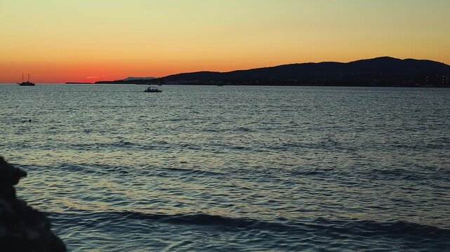 Sunset over calm water with boats sailing, silhouetted trees framing the scene, and distant hills creating a serene landscape in a coastal region