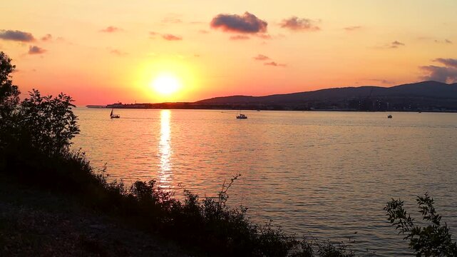 Sunset over calm water with boats sailing, silhouetted trees framing the scene, and distant hills creating a serene landscape in a coastal region