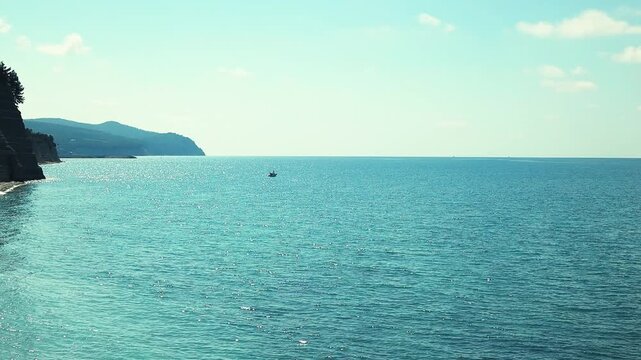 Small fishing boat glides through calm blue waters near a rocky coastline, with distant mountains and a clear sky enhancing the peaceful coastal landscape