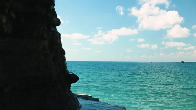 Coastal scene featuring turquoise ocean waves meeting rocky shore under a clear blue sky with fluffy clouds and a distant boat on the horizon