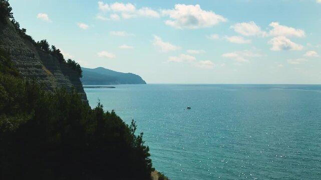 Small fishing boat glides through calm blue waters near a rocky coastline, with distant mountains and a clear sky enhancing the peaceful coastal landscape
