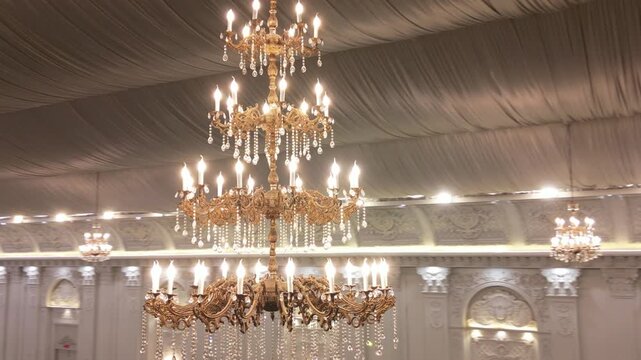 Close up of multi-tiered gold chandelier with lit candle-shaped bulbs and hanging crystal pieces inside a marquee hall captured in a drone orbit shot.