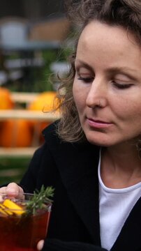 Woman savors aroma of amber cocktail, lifting its garnished glass near her face before lowering it. Relaxed bar guest pauses outdoors, appreciating drinks herbal garnish as she brings glass near her